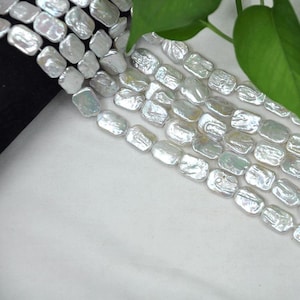 Puede incluir: Primer plano de hileras de perlas de agua dulce blancas, rectangulares. Las perlas tienen un brillo iridiscente sutil y están dispuestas en filas ordenadas. El fondo es una tela blanca suave, con un borde oscuro y una hoja verde.