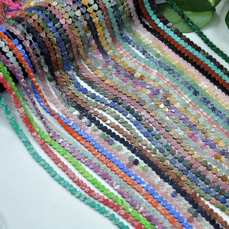 Puede incluir: Una colecci&oacute;n de cuentas de piedras preciosas en forma de coraz&oacute;n en varios colores, incluyendo rojo, verde, azul y morado. Las cuentas est&aacute;n ensartadas, creando hebras de componentes de joyer&iacute;a. La imagen muestra una variedad de texturas y tonos.