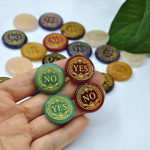 May include: A selection of round game pieces in various colours, with the words "YES" and "NO" in gold lettering. The pieces are green, red, blue, and gold. Some pieces are held in a hand.
