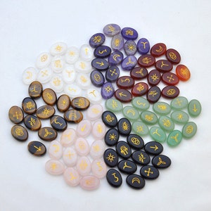 May include: A collection of oval-shaped stones with gold symbols, arranged in a circle. The stones are in seven different colors: white, brown, pink, black, green, purple, and red.