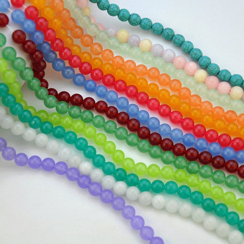 Puede incluir: Una colecci&oacute;n de 14 hebras de cuentas redondas, opacas, con acabado mate, en varios colores, incluyendo turquesa, azul claro, lavanda, blanco, verde, naranja y rojo. Las cuentas est&aacute;n enhebradas sobre un fondo blanco.