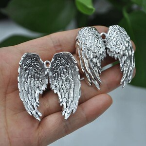 May include: Two silver-colored angel wing charms. The charms are detailed with feather-like textures and have a loop at the top for attaching to jewelry or other crafts. The wings are displayed against a blurred green background.