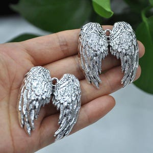 May include: Silver-colored angel wing charms. The charms are detailed with feather textures and have a loop at the top for attaching to jewelry or other crafts. The wings are displayed against a blurred background.