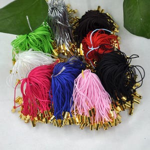 May include: A selection of colourful elastic cords with gold-coloured metal crimp ends. The cords are in various colours, including green, white, pink, red, blue, black, and silver. These cords are likely used for jewellery making or crafting.