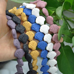 May include: Strands of colorful, porous lava rock beads in various shapes and colors, including purple, black, yellow, blue, white, and pink. The beads are held in a hand, showcasing their texture and natural appearance. Ideal for jewelry making.