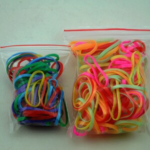 Pu&ograve; includere: Due sacchetti di plastica trasparente pieni di elastici colorati. Il sacchetto a sinistra contiene una variet&agrave; di colori, tra cui rosso, blu, verde, giallo e viola. Il sacchetto a destra contiene una variet&agrave; di colori vivaci, tra cui rosa, arancione, giallo e verde.