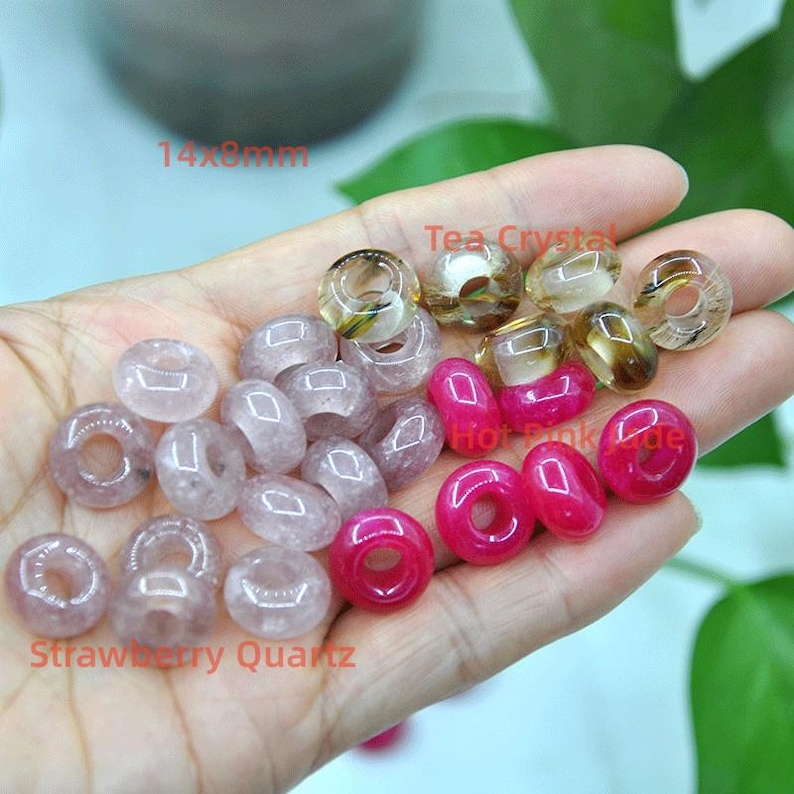 May include: A collection of 14mm x 8mm donut-shaped beads in various colors, including pink, red, and clear. The beads are made of natural stone and have a smooth, polished finish. The text "Strawberry Quartz" is visible in the image.