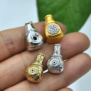 May include: Four small, decorative bottle-shaped beads in gold and silver tones. Each bead features intricate embossed designs and a hole for stringing. The beads are likely for jewelry making or other craft projects.