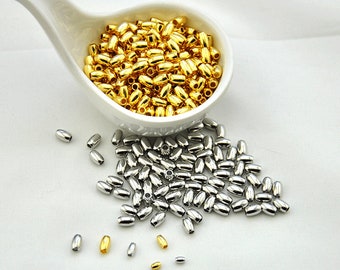 May include: A white ceramic spoon filled with gold-colored metal beads. A pile of silver-colored metal beads are scattered on a white surface.