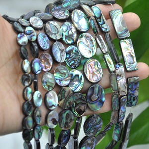 Könnte beinhalten: Nahaufnahme von Strängen aus Abalone-Muschelkernperlen in ovalen und rechteckigen Formen. Die Perlen weisen einen schimmernden, irisierenden Effekt mit Farben wie Blau, Grün und Lila auf. Die Perlen sind aufgefädelt und bereit für die Schmuckherstellung.