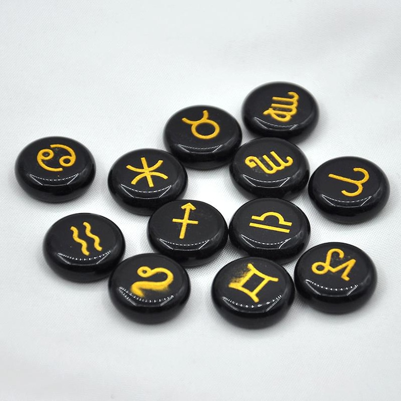 May include: Twelve black round stones with gold zodiac symbols. The symbols include Aries, Taurus, Gemini, Cancer, Leo, Virgo, Libra, Scorpio, Sagittarius, Capricorn, Aquarius, and Pisces.