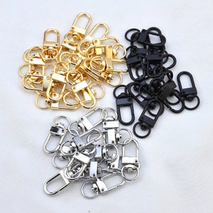 May include: A collection of gold, silver, and black metal swivel clasps. The clasps are in a pile and are all the same size and shape.