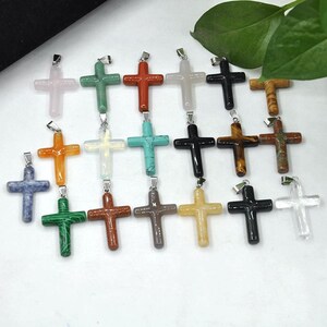 May include: A selection of gemstone cross pendants in diverse colours, such as pink, green, orange, and black. Each cross features a silver-coloured bail for attaching to a chain. The crosses are made from various materials.