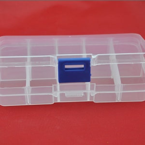 May include: A clear plastic organizer box with multiple compartments. The box has a blue latch and is designed for storing small items. The dividers create separate sections for organization. The box is on a red background.