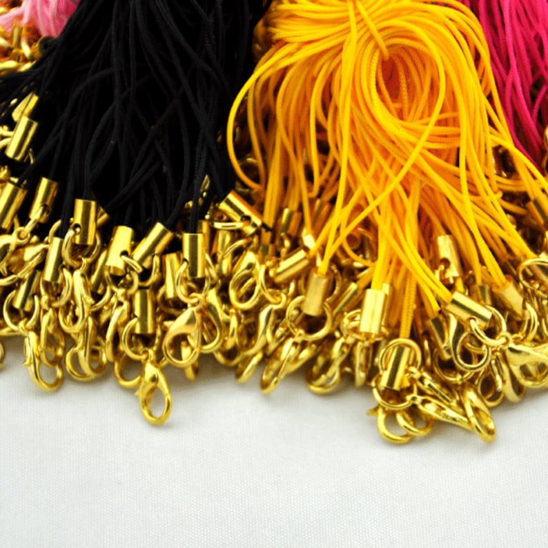 May include: A close-up of a pile of gold-colored lobster clasps attached to black, yellow, and pink cords. The cords are used for making phone charms, keychains, or other accessories.