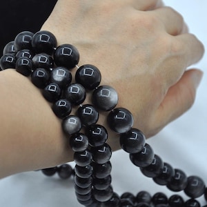 Könnte beinhalten: Ein Armband aus schwarzen Edelsteinen mit silbernem Schimmer. Das Armband besteht aus runden Perlen und wird am Handgelenk getragen.