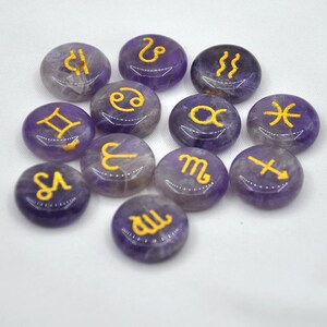 May include: Twelve amethyst gemstone tumbled stones with gold zodiac symbols. The stones are arranged in a circle on a white background.
