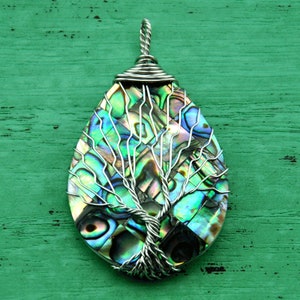 Op de afbeelding: Een zilveren draadgewikkelde hanger met een boom des levens ontwerp met iriserende abalone schelp inlay. De hanger is traanvormig en heeft een zilveren lus.