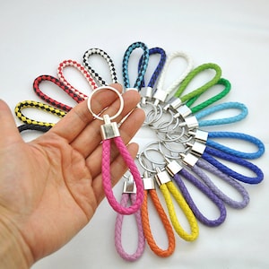 Op de afbeelding: Een verzameling kleurrijke gevlochten leren sleutelhangers met zilveren sleutelringen. De sleutelhangers zijn in een cirkel gerangschikt met de roze sleutelhanger in het midden.