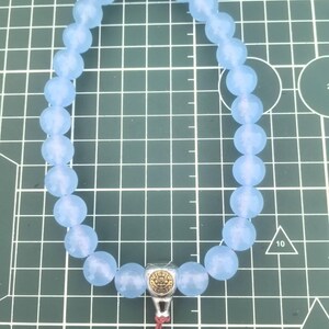 May include: A light blue beaded bracelet with a silver-colored charm and a red tassel. The bracelet features round, faceted beads. The charm has a gold-colored design. The bracelet is on a green grid background.