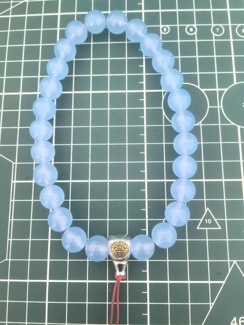 May include: A light blue beaded bracelet with a silver-colored charm and a red tassel. The bracelet features round, faceted beads. The charm has a gold-colored design. The bracelet is on a green grid background.