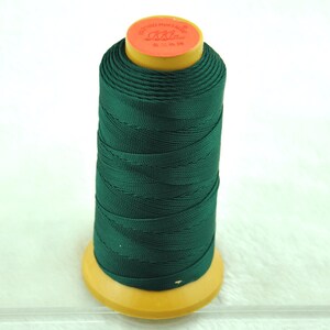May include: A spool of dark green thread with a yellow base and top. The top of the spool has red and gold text. The thread is tightly wound, creating a cylindrical shape. The thread is likely used for sewing or crafting.