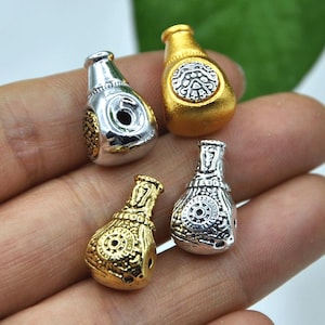 May include: Four small, decorative bottle-shaped beads in gold and silver tones. Each bead has intricate embossed designs and a small hole for stringing. The beads are held in a hand, showcasing their size and detail.
