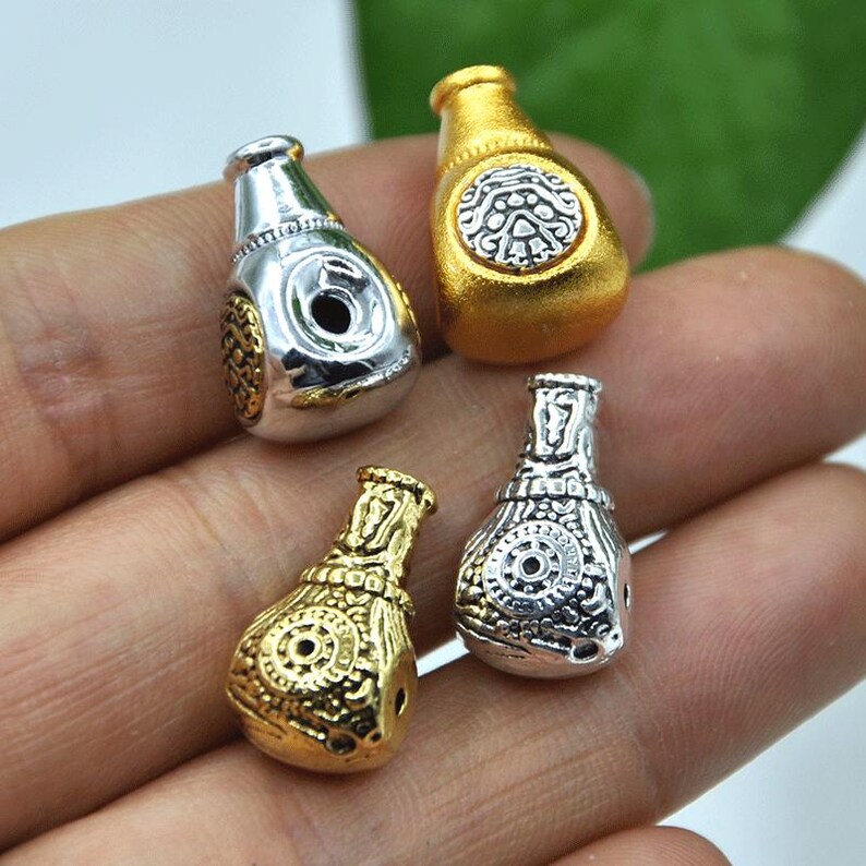 May include: Four small, decorative bottle-shaped beads in gold and silver tones. Each bead has intricate embossed designs and a small hole for stringing. The beads are held in a hand, showcasing their size and detail.