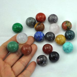 Peut inclure: Une collection de 18 sphères de pierres précieuses lisses et polies dans différentes couleurs, notamment le vert, le rouge, le bleu, le brun, le blanc et le rose. Les sphères sont disposées sur une main.