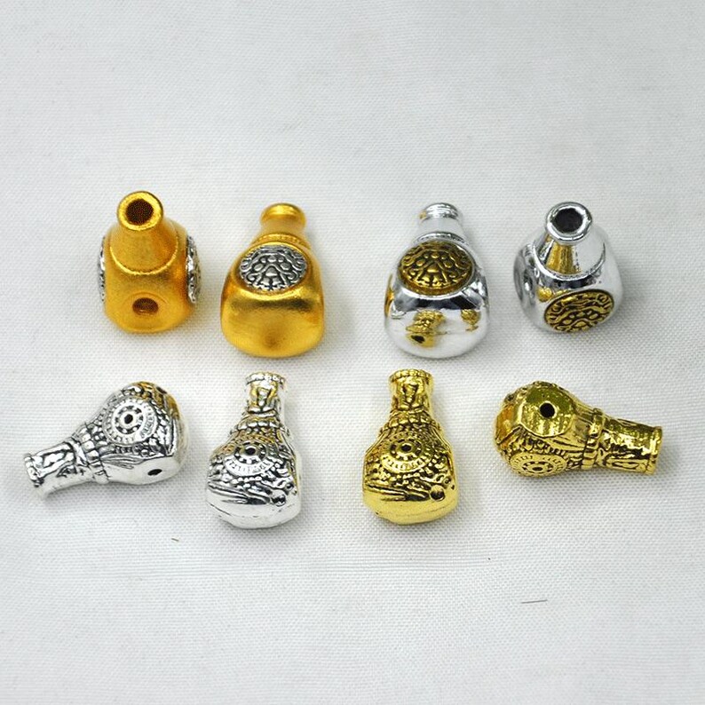 May include: A collection of eight decorative metal beads in gold and silver tones. The beads are shaped like small bottles with intricate embossed designs. Some beads have a matte finish, while others are shiny.