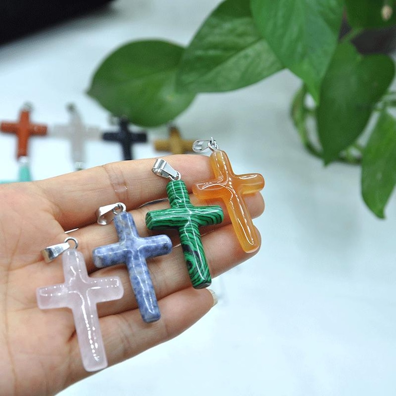 May include: A selection of gemstone cross pendants in shades of pink, blue, green, and orange. Each cross features a silver-coloured bail for attaching to a chain. The crosses are held in a hand, with green foliage in the background.