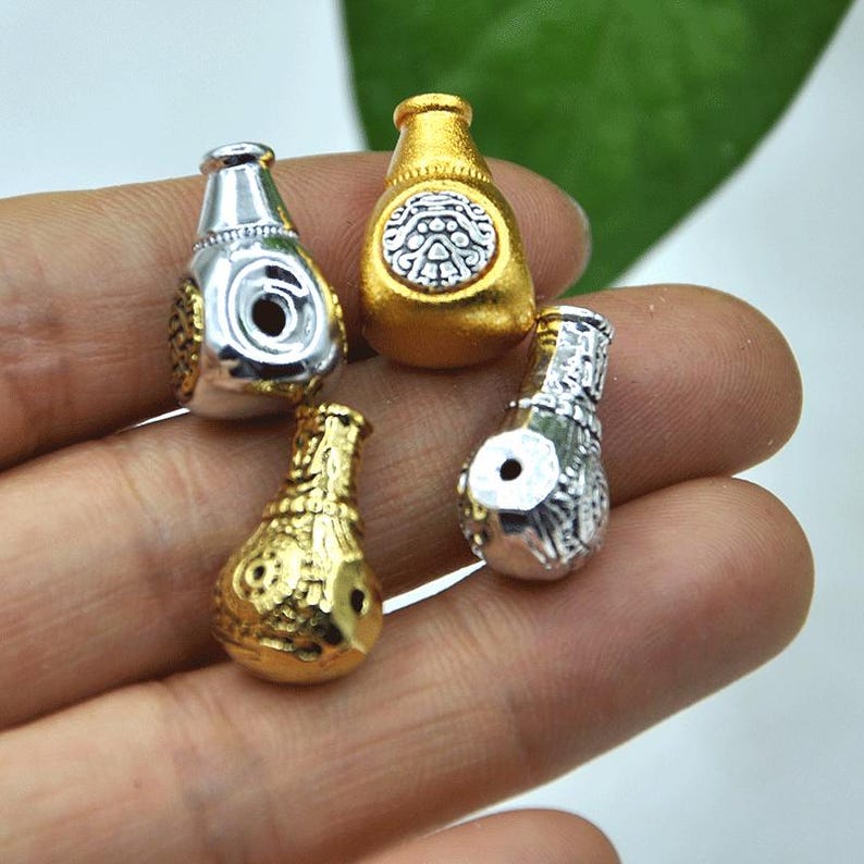 May include: Four small, decorative bottle-shaped beads in gold and silver tones. The beads have intricate patterns and a hole for stringing. The gold beads have a matte finish, while the silver beads are shiny.