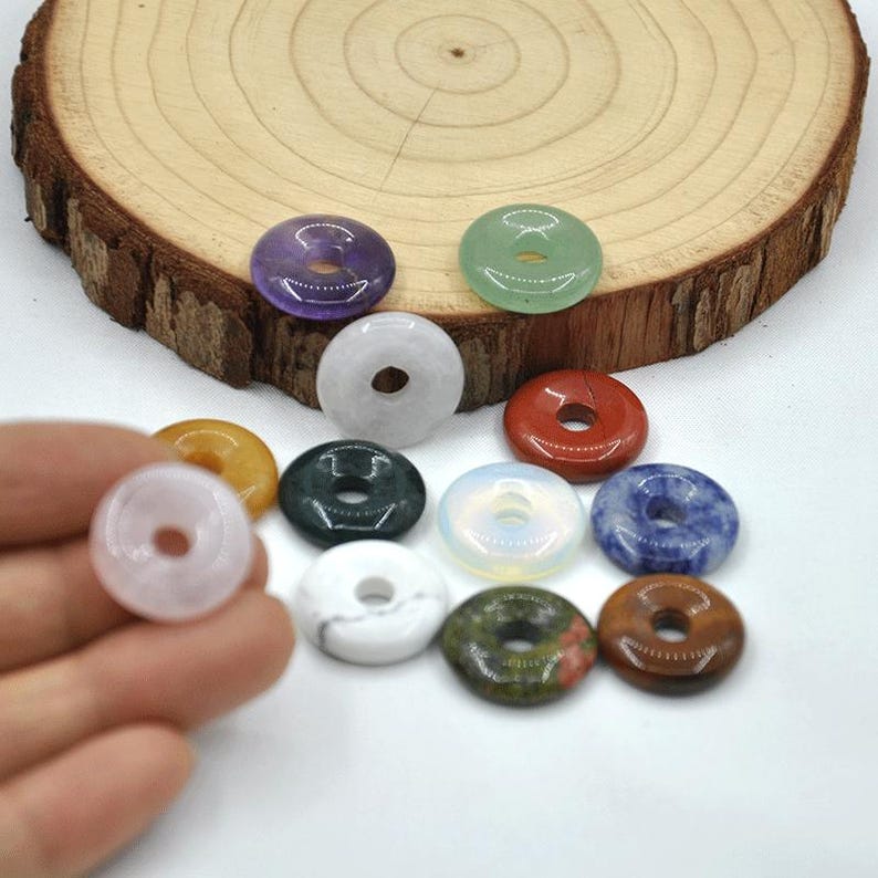 Pode incluir: Conjunto de 12 contas de pedras preciosas em forma de rosquinha em v&aacute;rias cores, incluindo branco, verde, vermelho, azul, marrom e roxo. As contas est&atilde;o dispostas sobre uma superf&iacute;cie de madeira.