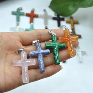 May include: A selection of gemstone cross pendants in shades of pink, blue, green, and orange. Each cross features a silver-coloured bail for attaching to a chain. The crosses are crafted from polished stone.