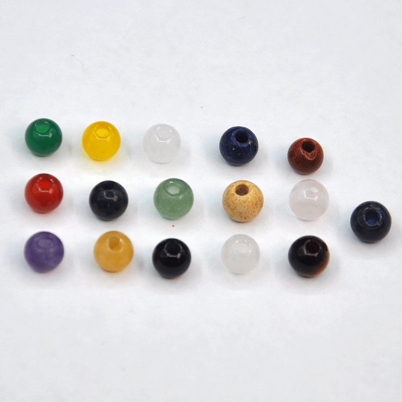 May include: Assortment of 15 round gemstone beads in various colors, including green, yellow, white, blue, red, black, purple, and brown. The beads are arranged in rows on a white surface.
