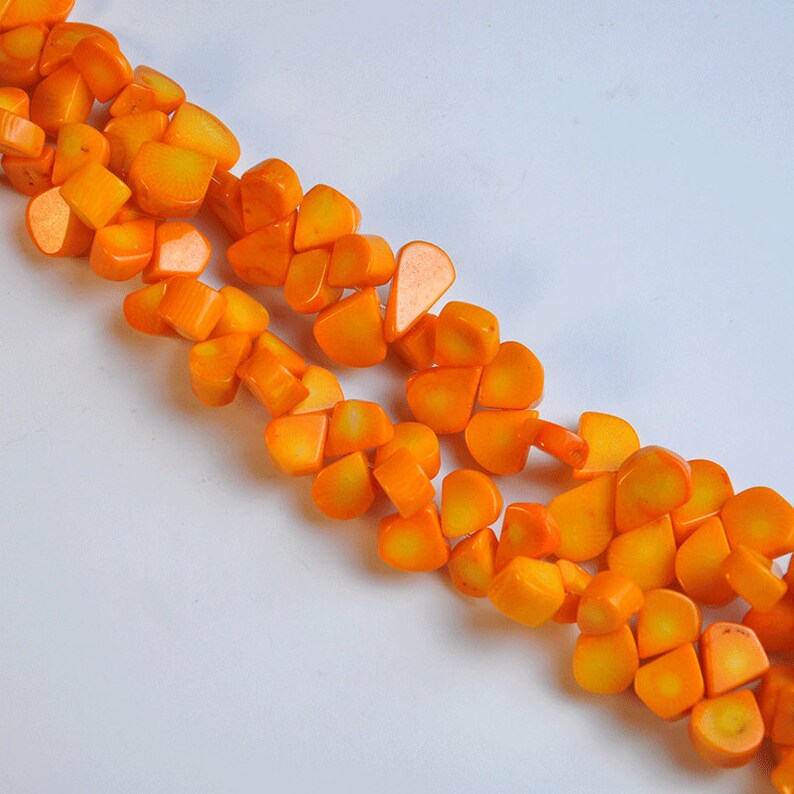 May include: Close-up of a necklace made of bright orange, irregularly shaped beads. The beads have a glossy finish and are strung together, creating a vibrant and eye-catching accessory. The necklace is set against a white background.