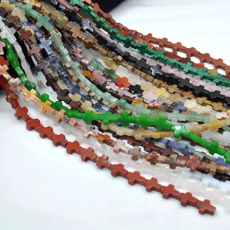 May include: A strand of gemstone beads in various colors, each bead is shaped like a cross. The beads are strung together on a clear thread. The beads are made of natural stone and have a variety of patterns and textures.