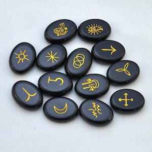May include: A set of 12 black oval-shaped stones with gold symbols etched on them. The symbols include a sun, a star, a crescent moon, a cross, an arrow, a spiral, an eye, a tree, a wave, a circle, a letter T, and a letter Y.