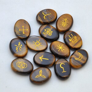 May include: A set of 12 tiger's eye stones, each with a different gold symbol etched on the surface. The stones are oval shaped and have a brown and gold color.