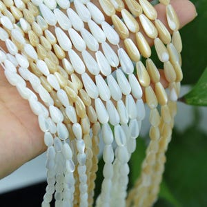 May include: Multiple strands of teardrop-shaped beads made from polished mother of pearl. The beads feature a smooth, iridescent finish with a color palette alternating between creamy off-white and soft beige tones, strung on thin white thread.
