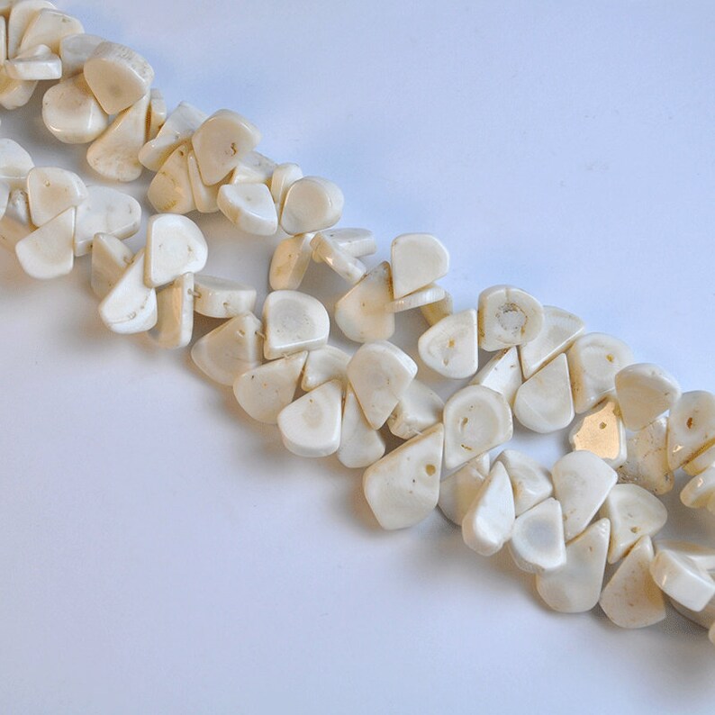 May include: Close-up of two strands of off-white, irregular-shaped beads. The beads have a polished, slightly glossy appearance and are strung together. The background is a plain white surface. These beads are suitable for jewelry making.