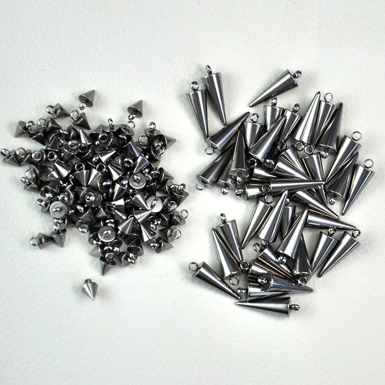 Puede incluir: Una gran cantidad de tachuelas de metal plateado en forma de pir&aacute;mide con un bucle en la parte superior. Las tachuelas est&aacute;n esparcidas sobre una superficie blanca.