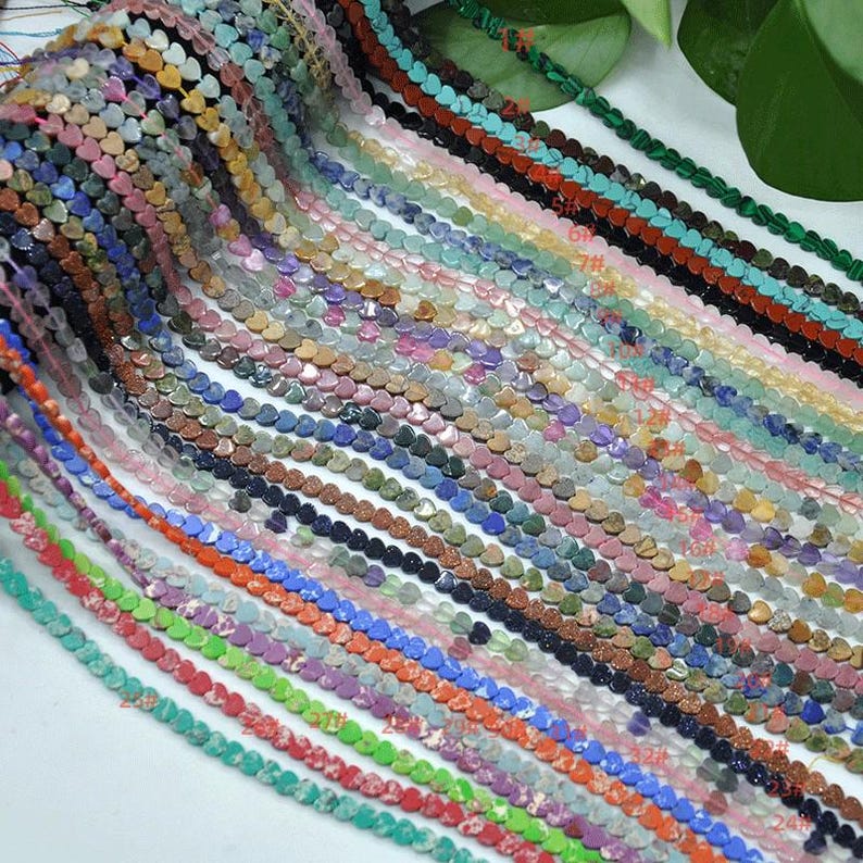 Puede incluir: Una colecci&oacute;n de cuentas en forma de coraz&oacute;n en varios colores, incluyendo rosa, azul, verde y marr&oacute;n. Las cuentas est&aacute;n ensartadas, creando m&uacute;ltiples hebras. La imagen muestra una variedad de cuentas de piedras preciosas, perfectas para la creaci&oacute;n de joyas.