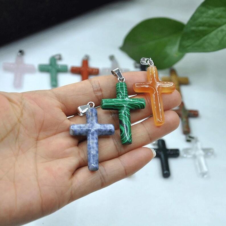 May include: A selection of gemstone cross pendants in various colours, including blue, green, orange, and clear. Each cross has a silver-tone bail for attaching to a chain. The crosses are made of different types of stone.