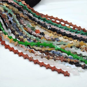 May include: A strand of colorful gemstone beads in the shape of crosses. The beads are made of various natural stones, including green, red, brown, blue, and white.