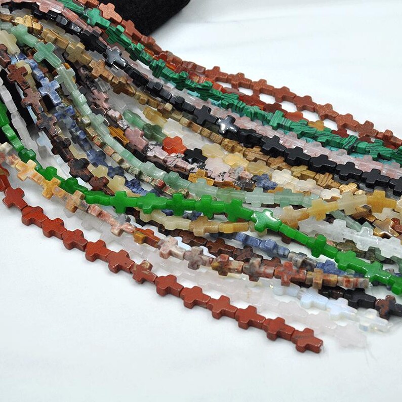 May include: A strand of colorful gemstone beads in the shape of crosses. The beads are made of various natural stones, including green, red, brown, blue, and white.
