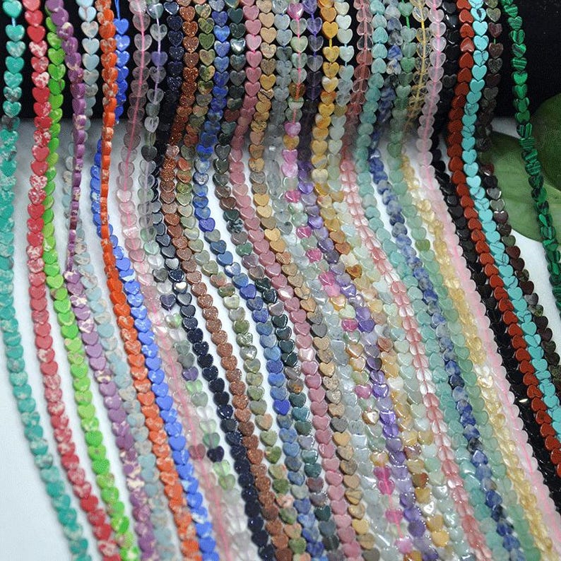 Puede incluir: Una colecci&oacute;n de cuentas en forma de coraz&oacute;n en varios colores, incluyendo rojo, verde, azul, morado y marr&oacute;n. Las cuentas est&aacute;n ensartadas, creando hebras de componentes de joyer&iacute;a coloridos. La imagen muestra una variedad de cuentas de piedras preciosas.