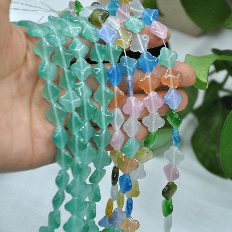 Puede incluir: Hebras de cuentas de piedras preciosas de colores en varias formas, incluyendo tr&eacute;boles y diamantes. Las cuentas son en tonos de verde, azul, rosa, naranja y blanco. Las cuentas se sostienen en una mano, con un fondo borroso.