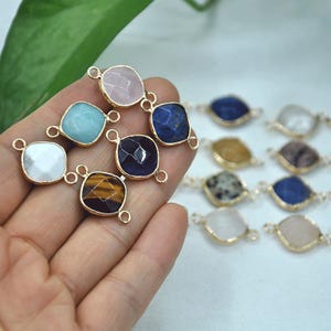 Puede incluir: Una colección de conectores de piedras preciosas en varios colores, incluyendo rosa, azul y marrón, con marcos de metal dorado. Las piedras preciosas son de forma cuadrada y tienen pequeños bucles para sujetar a otros componentes de joyería. Los conectores se muestran sobre un fondo suave.