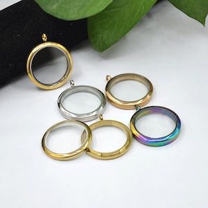 Può includere: Una collezione di ciondoli rotondi in vetro trasparente, in varie finiture metalliche, tra cui oro, argento e iridescente. Ogni ciondolo ha un piccolo anello per attaccarlo a una catena. I ciondoli sono esposti su una superficie bianca.